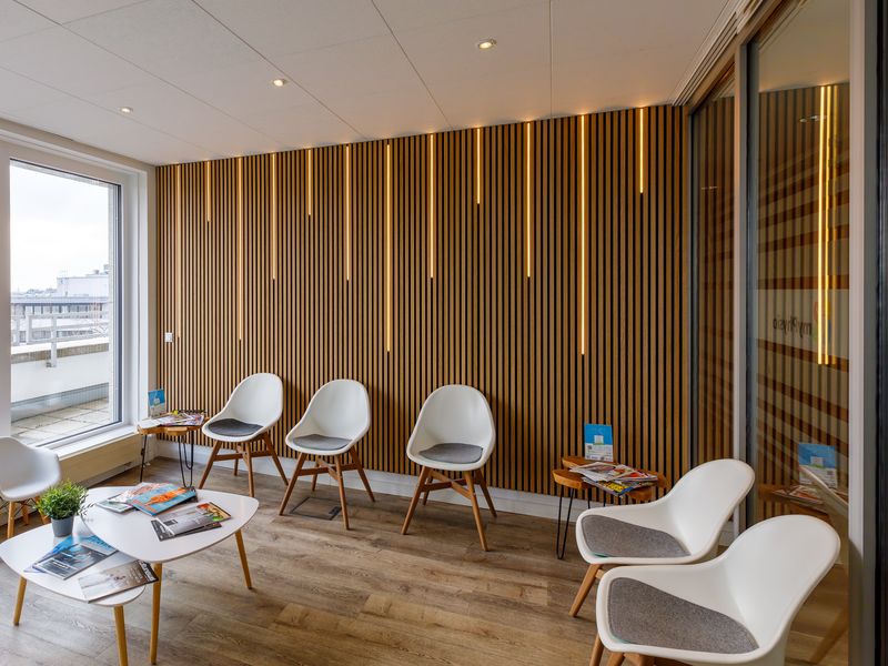 Wartebereich mit weißen Schalenstühlen vor einer beleuchteten Holzlamellenwand.