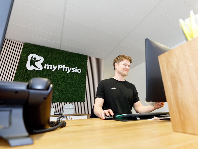 Empfangsmitarbeiter sitzt lächelnd am Computer, daneben die Mooswand mit dem myPhysio-Logo.