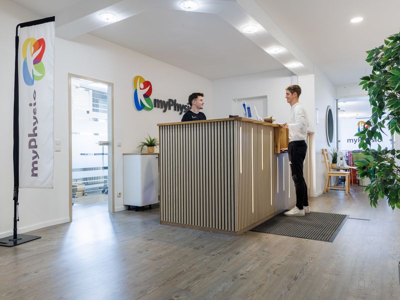 Empfang mit Holzlamellentresen und großem myPhysio-Logo, dahinter ein Mitarbeiter im Gespräch mit einem Patienten.