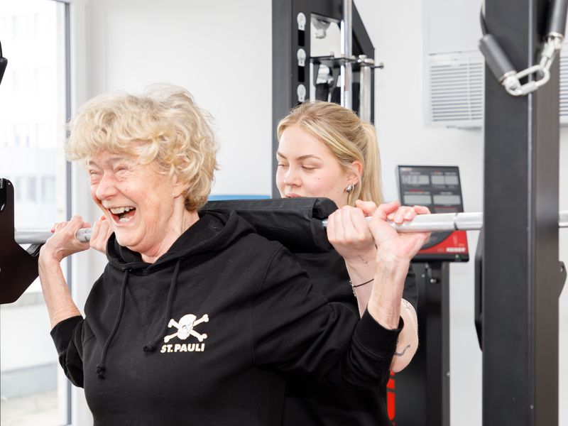 Ältere Patientin lacht beim Training mit der Langhantel, eine Therapeutin sichert sie von hinten.