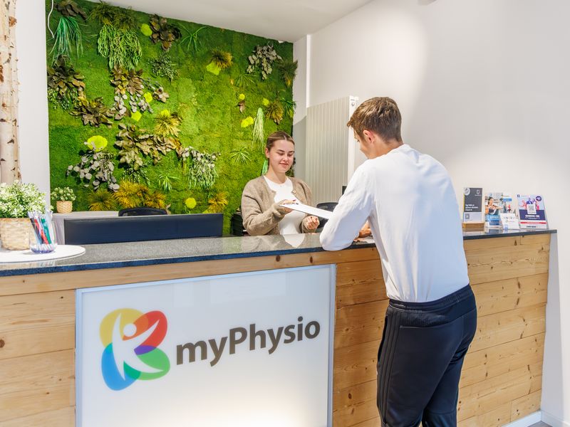 Empfang mit Mooswand und beleuchtetem Logo: Eine Mitarbeiterin reicht einem Patienten Unterlagen.