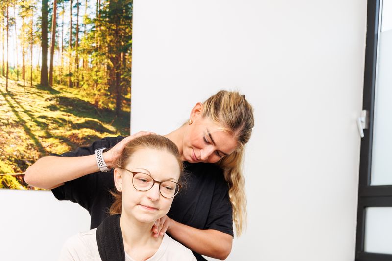 Therapeutin neigt den Kopf einer sitzenden Patientin zur Seite und fixiert mit der anderen Hand die Schulter.