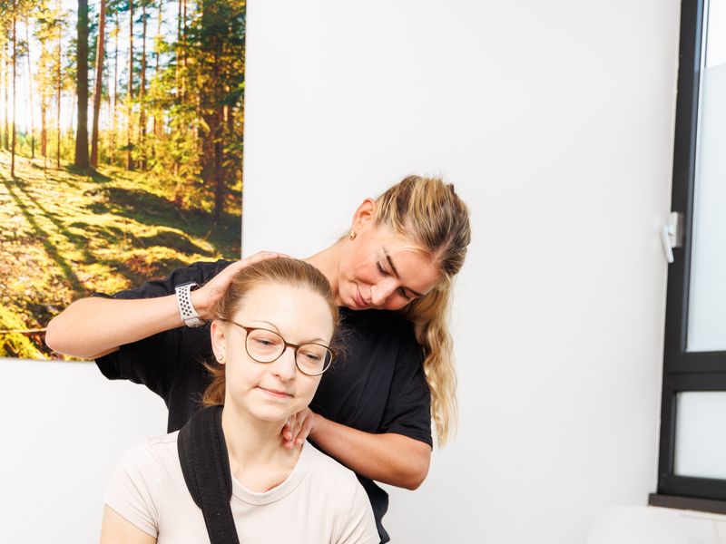 Therapeutin neigt den Kopf einer sitzenden Patientin zur Seite und fixiert mit der anderen Hand die Schulter.