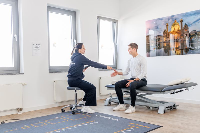 Therapeutin sitzt auf einem Hocker und fasst die Hand eines Patienten, der auf der Liege sitzt.