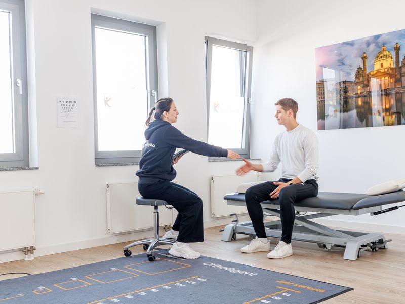 Therapeutin sitzt auf einem Hocker und fasst die Hand eines Patienten, der auf der Liege sitzt.