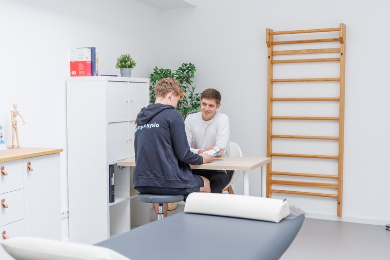 Therapeut zeigt einem Patienten am Tisch einen Flyer, eine Liege steht im Vordergrund.