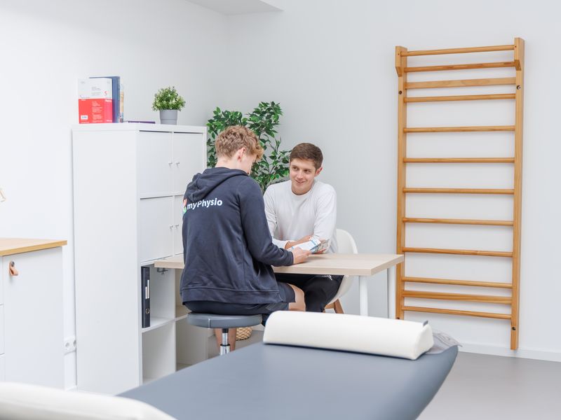 Therapeut zeigt einem Patienten am Tisch einen Flyer, eine Liege steht im Vordergrund.