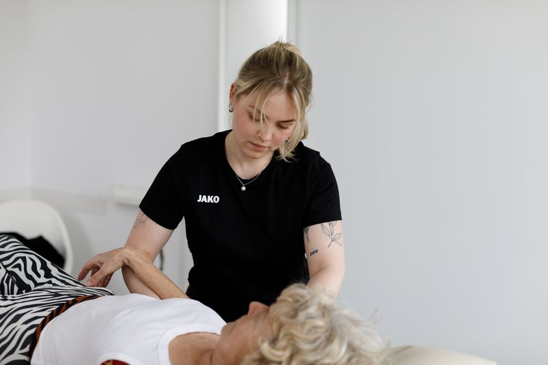 Therapeutin beugt sich über eine liegende Patientin und fasst ihren Arm.