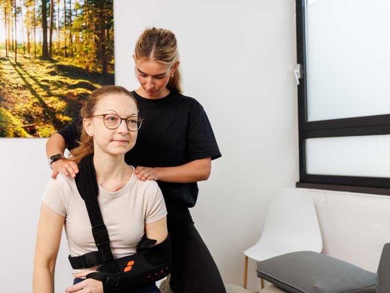 Therapeutin legt einer Patientin mit Schulterorthese beide Hände auf die Schultern.