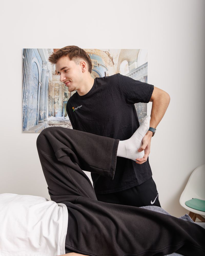 Therapeut beugt lächelnd das Bein eines liegenden Patienten im Hüftgelenk.