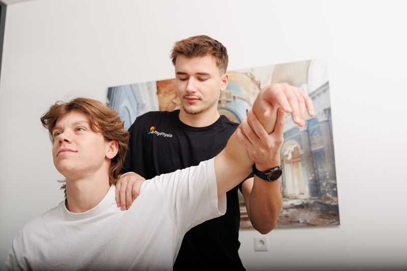 Therapeut hebt den Arm eines sitzenden Patienten an und fixiert mit der anderen Hand die Schulter.