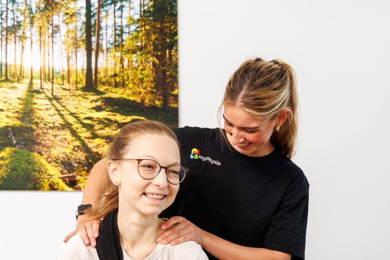 Patientin mit Schulterorthese lacht, die Therapeutin kniet hinter ihr und tastet Schultern und Nacken ab.